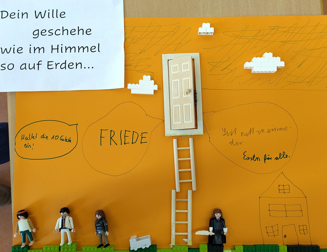 Ein Plakat mit dem Text "Dein Wille geschehe wie im Himmel so auf Erden". Darunter verschiedene Playmobil-Figuren und eine Treppe zu einer Himmelstür. Sprechblasen kommen aus der Tür, darauf steht: "Halte die 10 Gebote ein", "FRIEDE", "Seid nett zueinander", "Essen für alle".