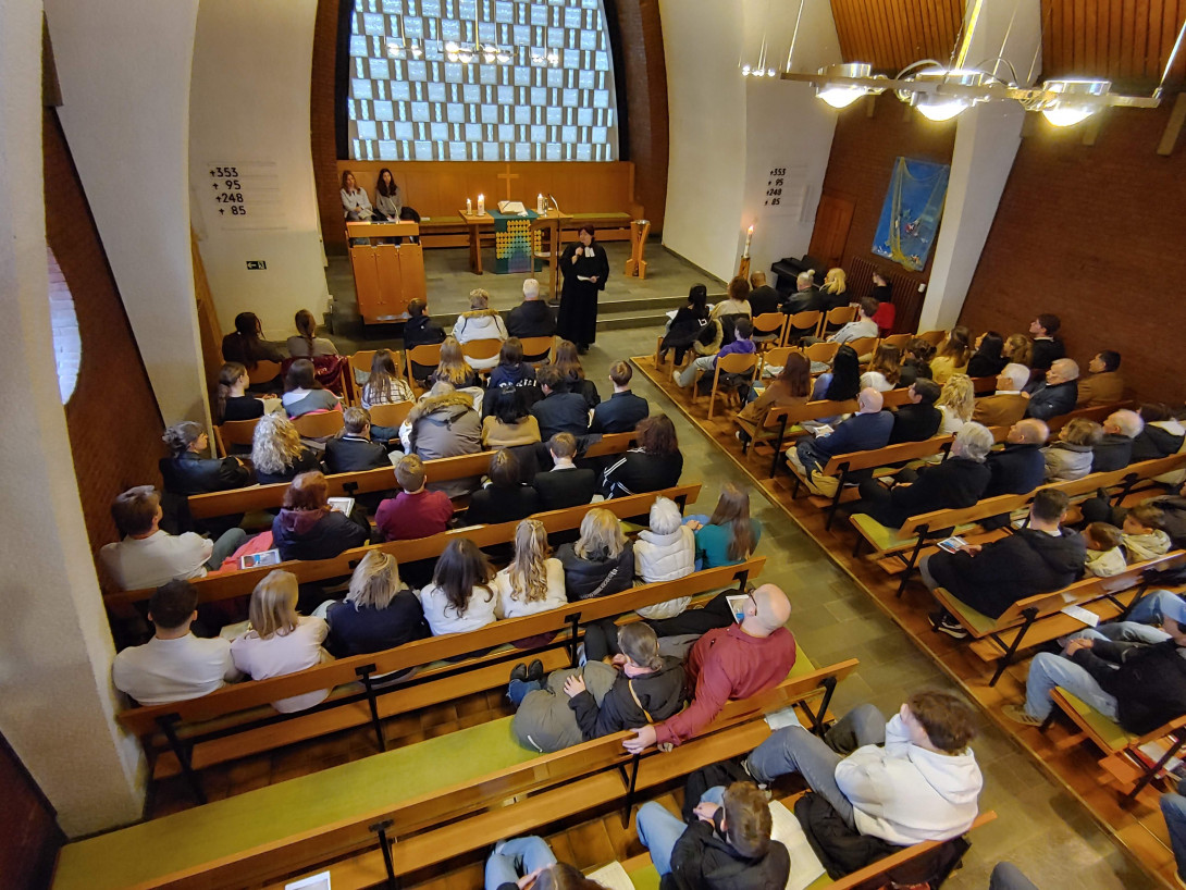 Blick in den Altar- und Kirchraum von oben. Viele Bänke sind voll besetzt. Eine Pfarrerin steht frontal.