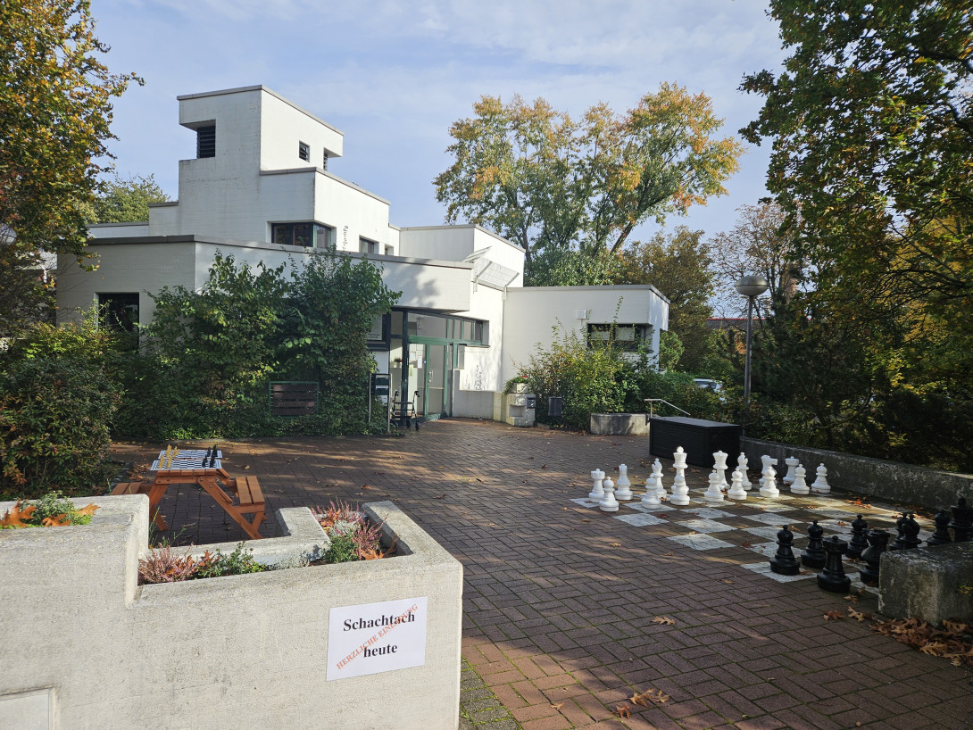 Im Vordergrund ein Schild an einer Mauer: "Schachtach". Dahinter ein Tisch auf dem ein Schachbrett liegt und daneben ein großes Boden-Schachbrett mit großen Spielfiguren. Im Hintergrund das Gemeindehaus Hochstraß.