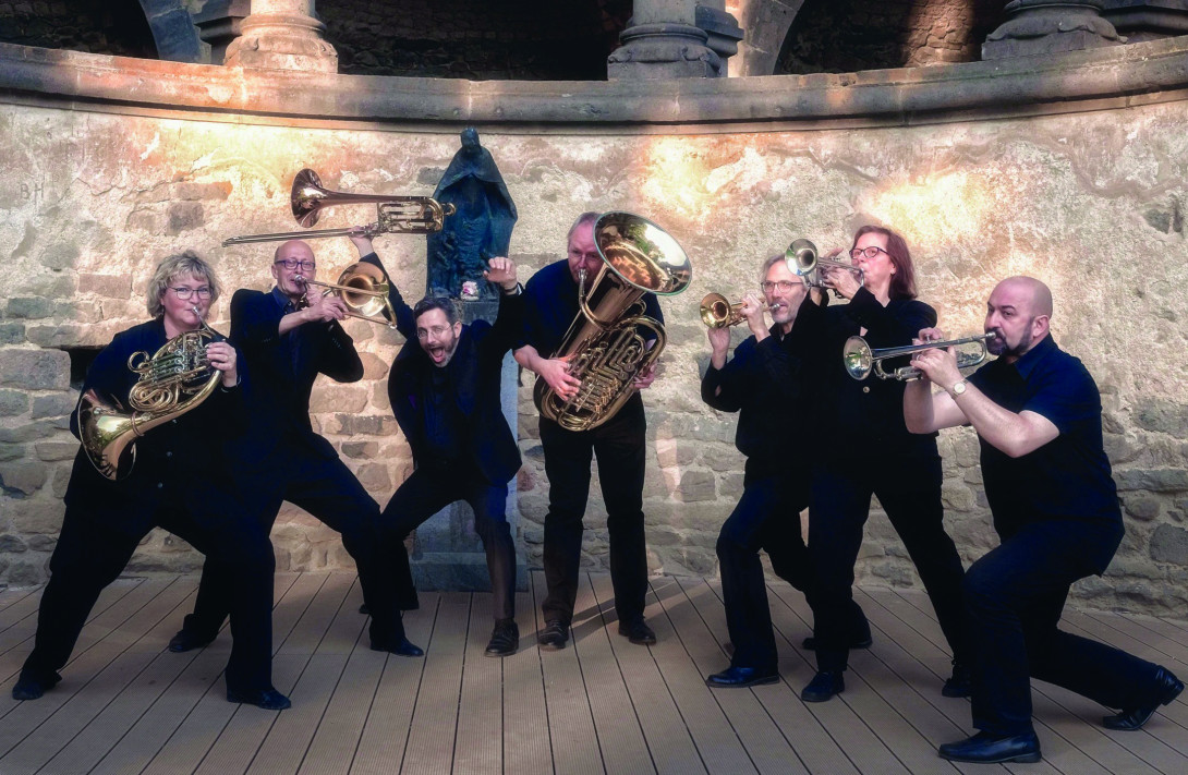 Band „Septomania Brass“. Die 7 Musiker in schwarzer Kleidung posieren in extrovertierten Posen mit Musikinstrumenten für ein Gruppenbild.