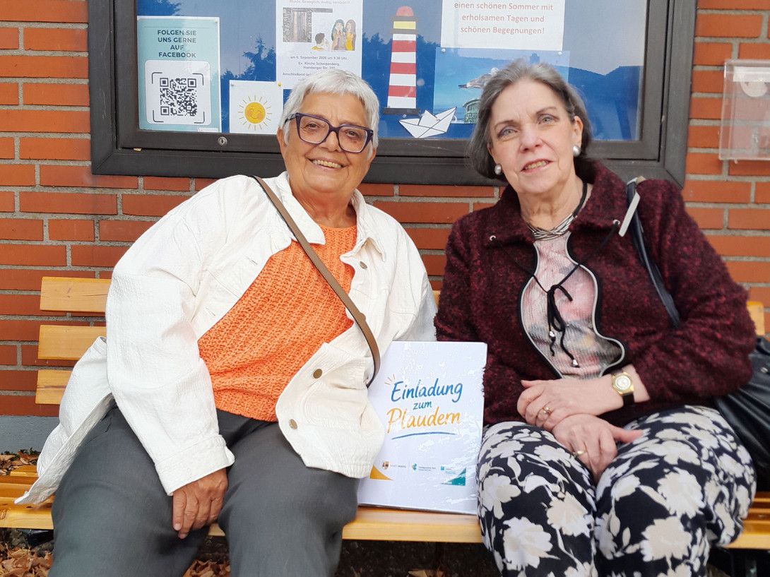 Zwei ältere Frauen sitzen auf einer Holzbank vor einem Schaukasten mit Plakaten, eine hält ein Schild mit der Aufschrift 'Einladung zum Plaudern'.