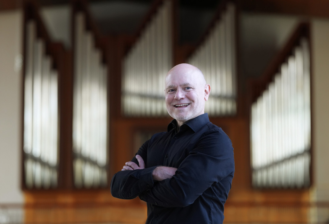 Künstler Patrick Gläser lächelnd und mit verschränkten Armen vor einer Orgel stehend.