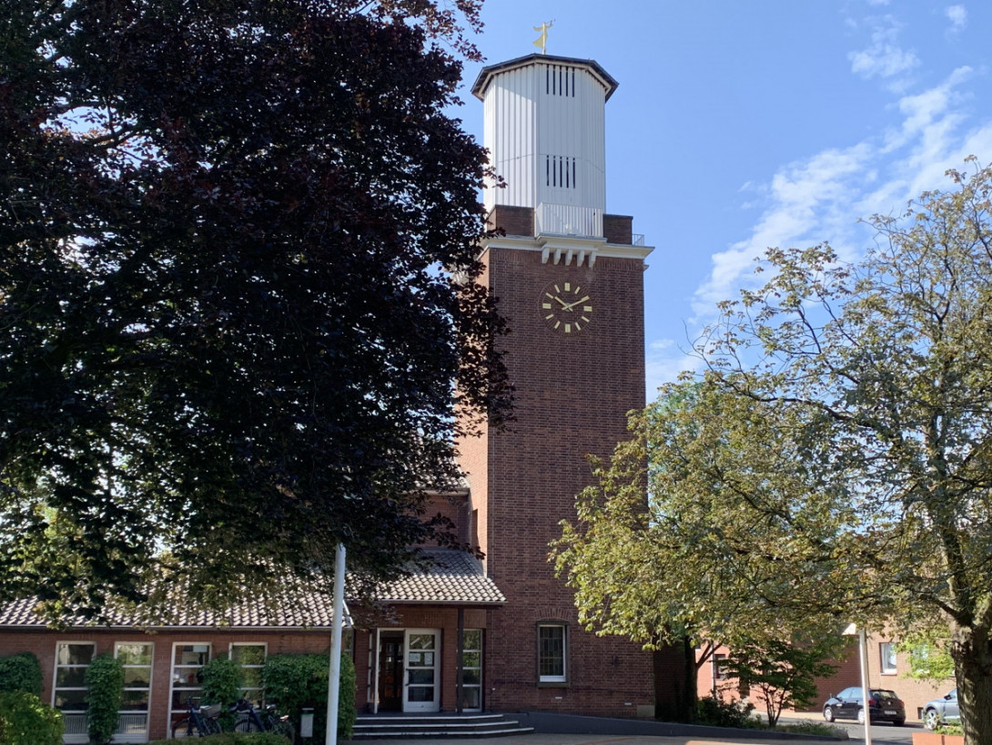 Kirchturm der Kirchengemeinde Moers-Asberg mit grünen Bäumen und blauem Himmel