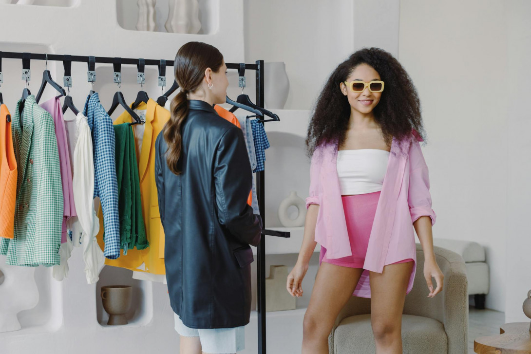 Zwei junge Frauen an einem Kleiderständer mit fesher Mode. Eine der Frauen trägt eine Sonnenbrille und schaut in die Kamera.