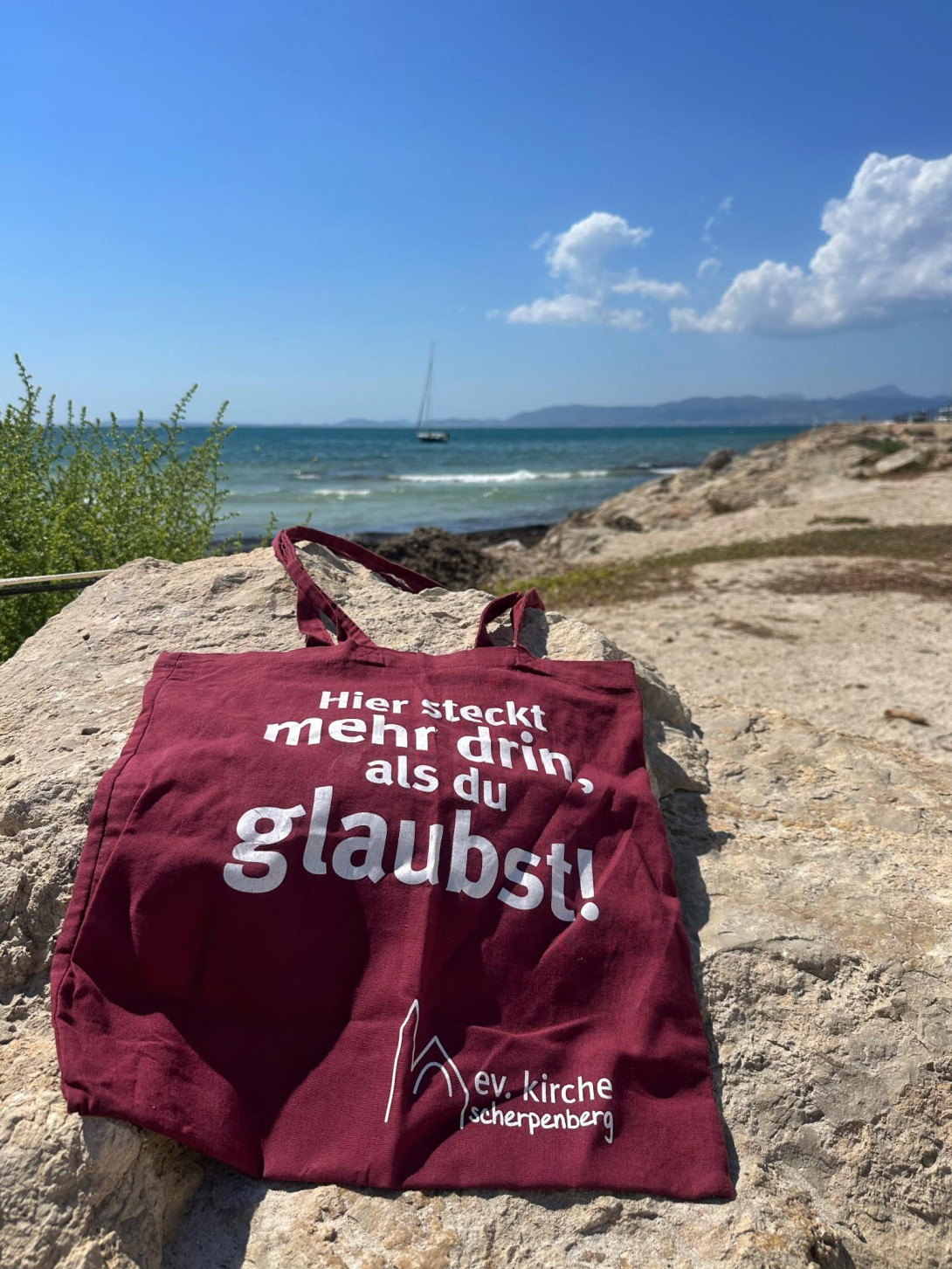 Gemeindetasche mit Aufdruck "Hier steckt mehr drin, als du glaubst!" liegt auf einem Felsen am Meer. Im Hintergrund die Meeresbrandung, ein Segelschiff und blauer Himmel mit wenigen Wolken.